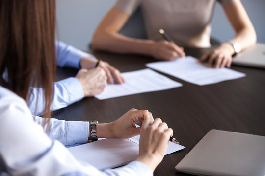Close up of business partners signing contracts during company briefing or team meeting, people sitting at shared table at conference, putting signature on documents or agreements at negotiations