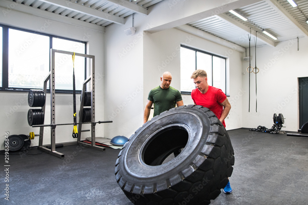 Poster Fit young man with a personal trainer in gym working out, moving tire.