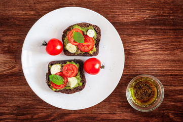 Sandwich toasts with tomatoes cherry, mozzarella, avocado, basil and olive oil. Top view on a dark wooden background. Flat lay.