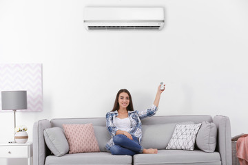 Young woman switching on air conditioner while sitting on sofa near white wall