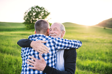 An adult hipster son with senior father on a walk in nature at sunset, hugging.