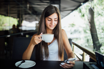 Charming woman with beautiful smile reading good news on mobile phone during rest in coffee shop. Happy Caucasian female watching her photo on cell telephone while relaxing in cafe during free time