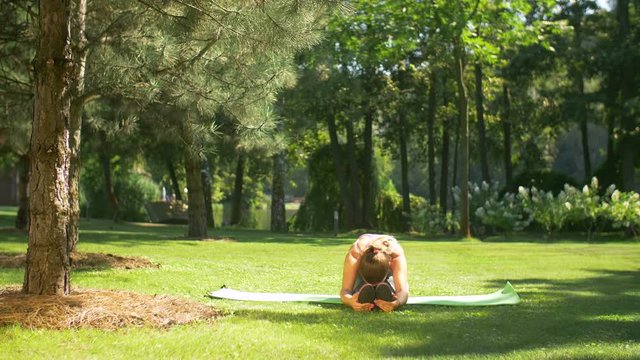 Beautiful fit woman in sportswear doing pilates training, seated forward bend pose on exercise mat in summer park. Fit active female stretching her body doing flexibility exercises and bending back.