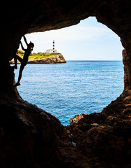 Silhouette of a female athlete climbing in a cave, Portocolom Lighthouse on a cliff in background, Mallorca, Spain.