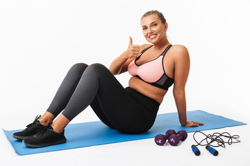 Pretty smiling girl with excess weight in sporty top and leggings sitting on yoga mat while happily showing thumb up gesture looking in camera over white background. Plus size model