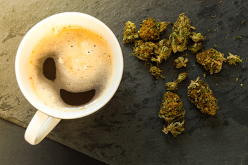 Delicious cup of coffee with a pile of high quality marijuana buds. Two drugs: cannabis and caffeine. Top view with background of black stone.