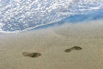traces of human feet on black sand near the water on the beach. Background texture.