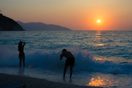 Silhouette man is taking pictures of a silhouette girl who is playing with waves at Myrtos beach in Kefalonia Greece. Sunset background