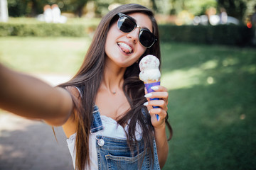Portrait of a girl wearing glasses with ice cream take selfie on the phone enjoying summer in the city park .
