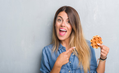 Young adult woman over grey grunge wall eating Belgium waffle very happy pointing with hand and finger