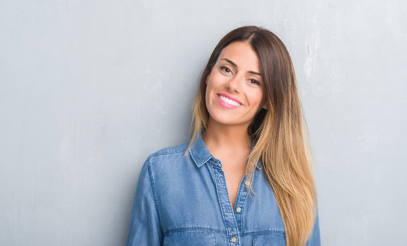 Young adult woman over grey grunge wall wearing denim outfit with a happy face standing and smiling with a confident smile showing teeth