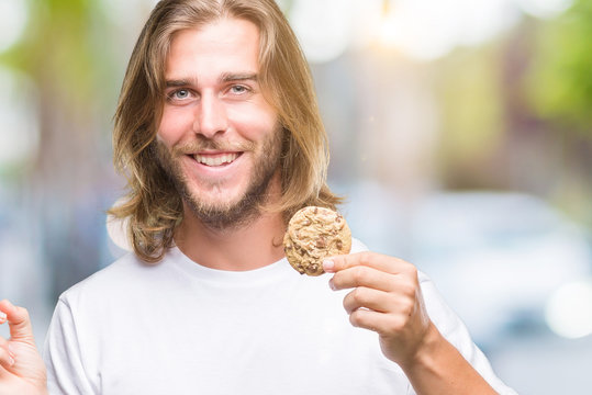 Young handsome man with long hair eating chocolate cooky over isolated background very happy pointing with hand and finger to the side