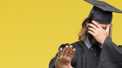 Young handsome graduated man with long hair over isolated background covering eyes with hands and...