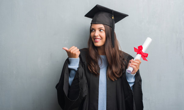 Young brunette woman over grunge grey wall wearing graduate uniform holding degree pointing and showing with thumb up to the side with happy face smiling