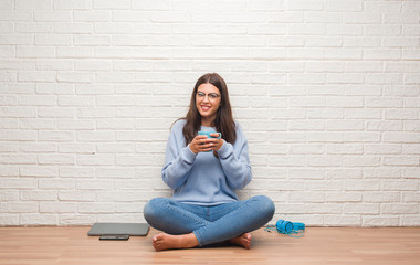 Young brunette woman sitting on the floor over white brick wall drinking coffee with a happy face standing and smiling with a confident smile showing teeth