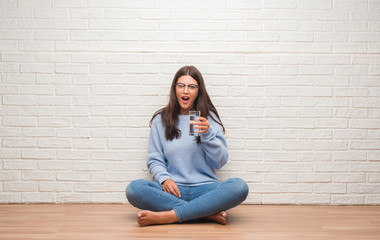 Young brunette woman sitting on the floor drinking glass of water scared in shock with a surprise face, afraid and excited with fear expression