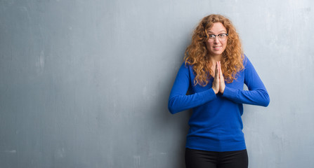 Young redhead woman over grey grunge wall praying with hands together asking for forgiveness smiling confident.