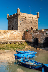 Eine Flotte blauer Fischerboote im Hafen von Essaouira. Sie können auch die Befestigungsanlagen und einen Turm der Zitadelle von Mogador sehen © Louis-Michel DESERT