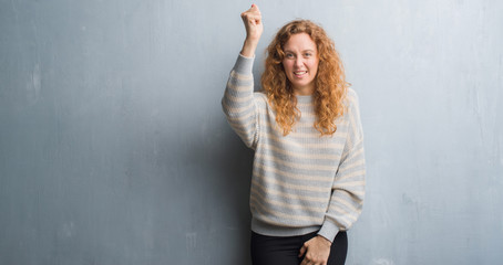 Young redhead woman over grey grunge wall angry and mad raising fist frustrated and furious while shouting with anger. Rage and aggressive concept.