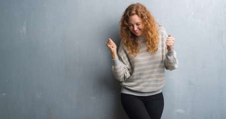 Young redhead woman over grey grunge wall very happy and excited doing winner gesture with arms raised, smiling and screaming for success. Celebration concept.