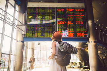Theme travel public transport. young woman standing with back in dress and hat behind backpack and camping equipment for sleeping, insulating mat looks schedule on scoreboard airport station © Elizaveta