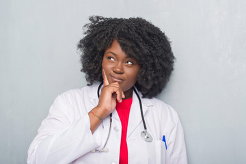 Young african american doctor woman over grey grunge wall with hand on chin thinking about question, pensive expression. Smiling with thoughtful face. Doubt concept.