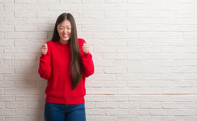 Young Chinese woman over brick wall excited for success with arms raised celebrating victory smiling. Winner concept.