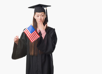 Young Chinese woman wearing graduate uniform holding United states of america flag cover mouth with hand shocked with shame for mistake, expression of fear, scared in silence, secret concept