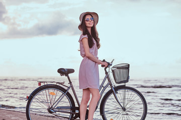 Naklejka premium Happy sensual girl in sunglasses and hat wearing dress walks with her bicycle on the beach against an amazing seaside background.