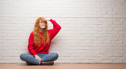 Young redhead woman sitting over brick wall very happy and smiling looking far away with hand over head. Searching concept.