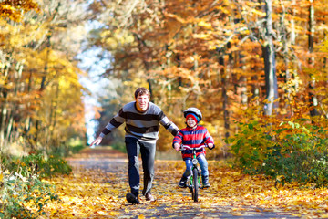 Fototapeta premium Little preschool kid boy and his father in autumn park with a bicycle. Dad teaching his son biking. Active family leisure. Child with helmet on bike. Safety, sports, leisure with kids concept..