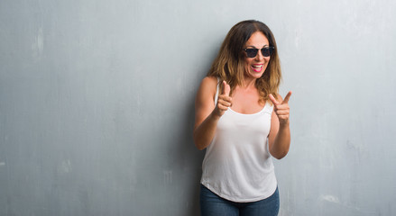 Middle age hispanic woman over grey wall wearing sunglasses pointing fingers to camera with happy and funny face. Good energy and vibes.