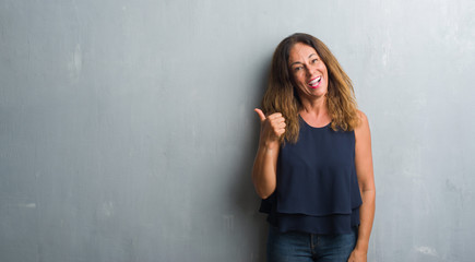 Middle age hispanic woman standing over grey grunge wall smiling with happy face looking and pointing to the side with thumb up.