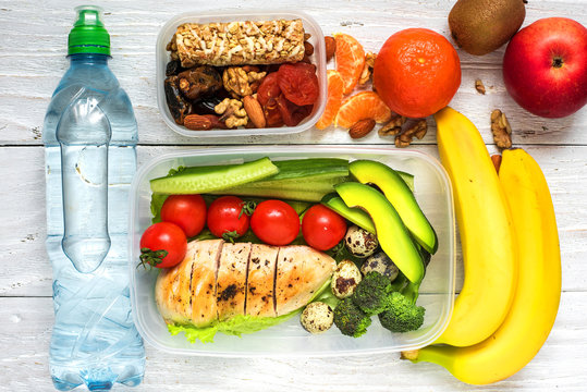 Healthy lunch boxes with chicken, fresh vegetables, fruits and nuts with bottle of water on white wooden background