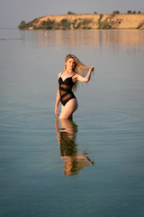 Girl in a black swimsuit with lush blond hair stands in sunny weather in the water