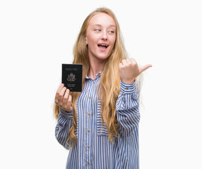 Blonde teenager woman holding passport of Unites States of America pointing and showing with thumb up to the side with happy face smiling