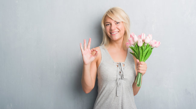 Caucasian adult woman over grey grunge wall holding pink flowers doing ok sign with fingers, excellent symbol
