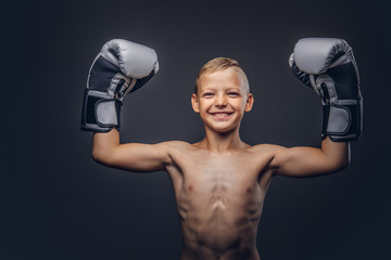 Cheerful shirtless little boy boxer with blonde hair wearing boxing gloves rejoices in a victory.