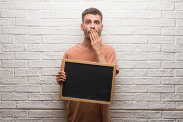 Young adult man over brick wall holding blackboard cover mouth with hand shocked with shame for mistake, expression of fear, scared in silence, secret concept