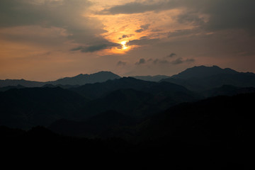 Obraz premium Sunset Abstract Graphic Resource. Panoramic Mountains, Clouds and Sun behind clouds in the shape of an orange lowing ball, opening in the clouds. Sign from heaven concept, calm mountains sundown.