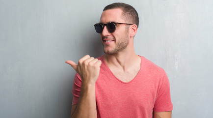 Young caucasian man over grey grunge wall wearing sunglasses pointing and showing with thumb up to...