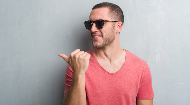 Young caucasian man over grey grunge wall wearing sunglasses pointing and showing with thumb up to the side with happy face smiling