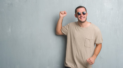 Young caucasian man over grey grunge wall wearing sunglasses angry and mad raising fist frustrated and furious while shouting with anger. Rage and aggressive concept.