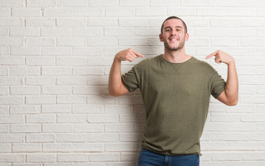 Young caucasian man standing over white brick wall looking confident with smile on face, pointing oneself with fingers proud and happy.