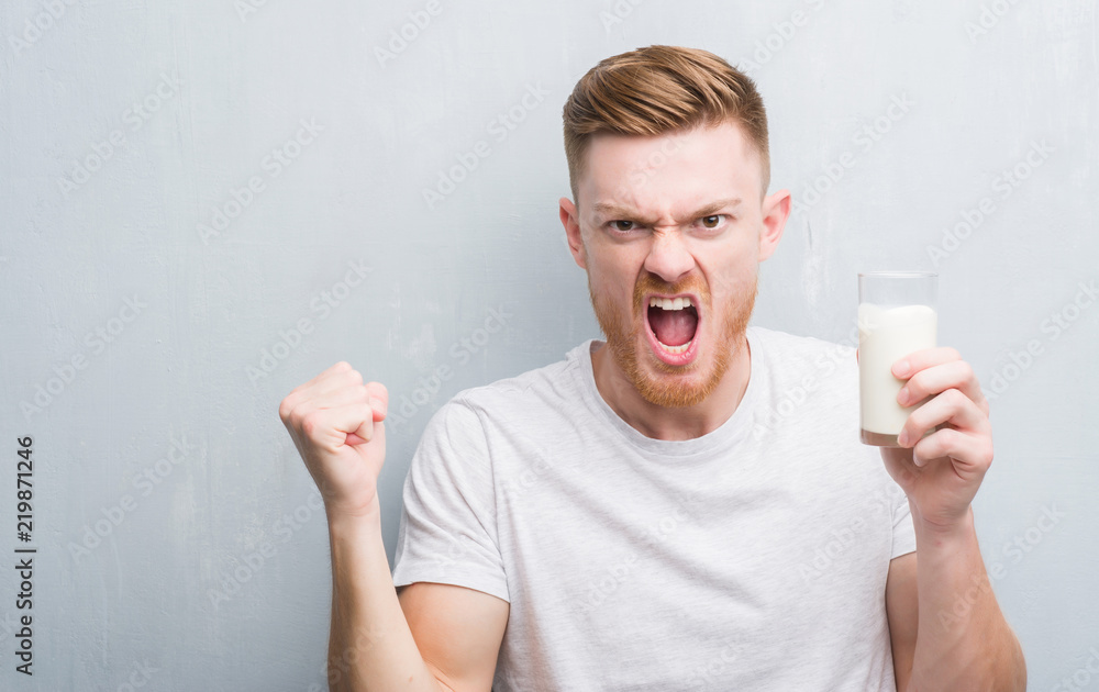 Wall mural Young redhead man over grey grunge wall drinking a glass of milk annoyed and frustrated shouting with anger, crazy and yelling with raised hand, anger concept