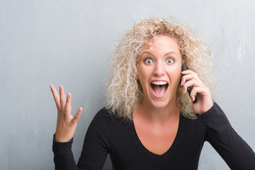 Young blonde woman over grunge grey background talking on the phone very happy and excited, winner expression celebrating victory screaming with big smile and raised hands