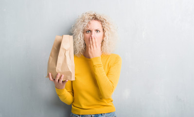 Young blonde woman over grunge grey background holding lunch paper bag cover mouth with hand...