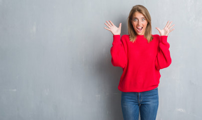 Beautiful young woman standing over grunge grey wall wearing winter sweater very happy and excited, winner expression celebrating victory screaming with big smile and raised hands