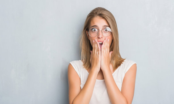 Beautiful young woman standing over grunge grey wall wearing glasses cover mouth with hand shocked with shame for mistake, expression of fear, scared in silence, secret concept