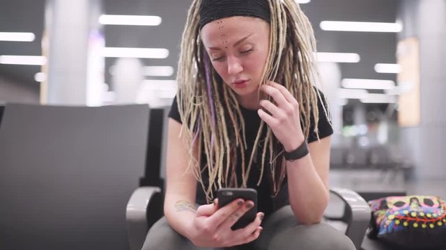 Attractive young woman blogger with dreadlocks using phone at the airport. Portrait of beautiful hipster woman